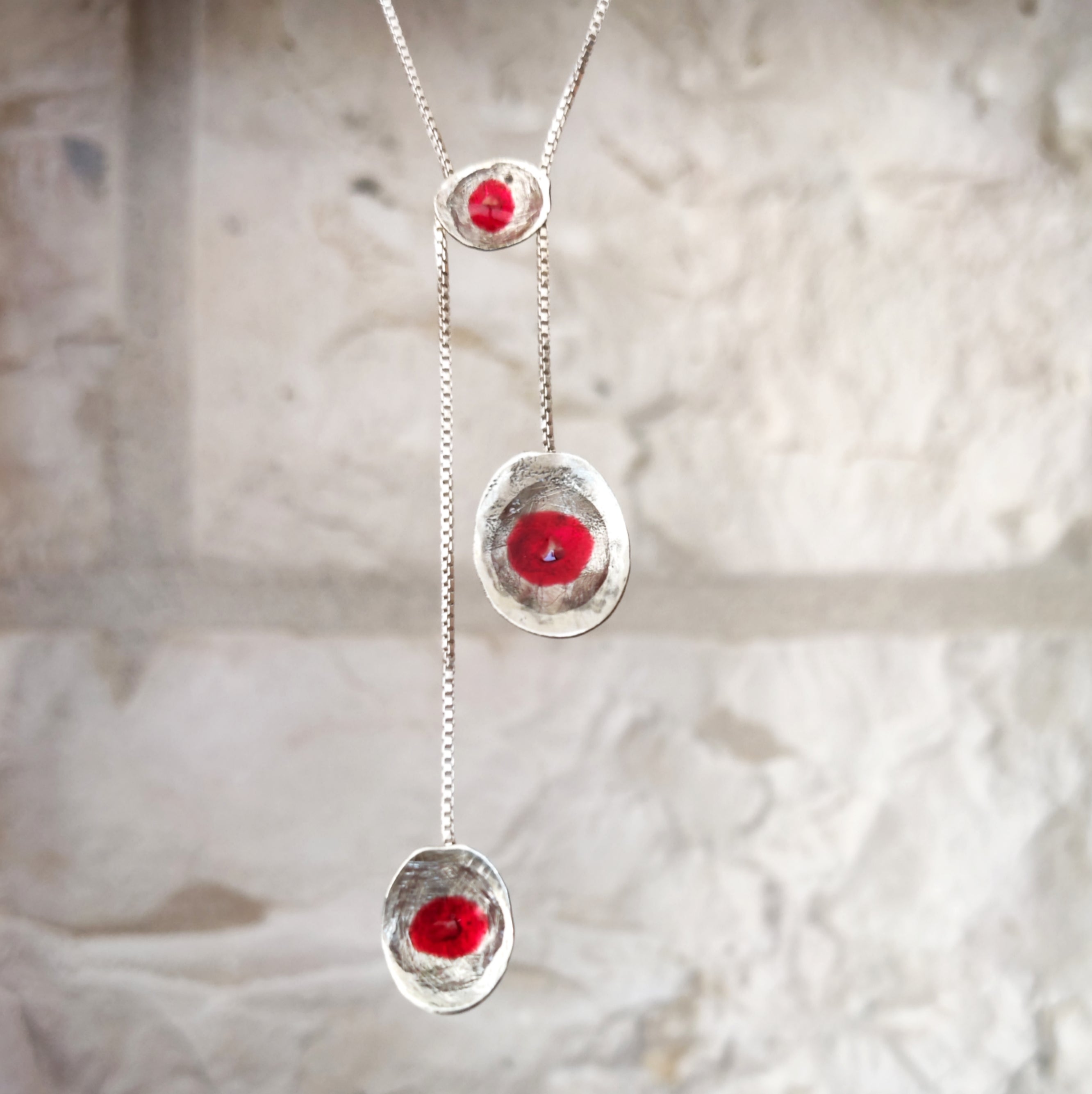A silver lariat-style necklace with three dangling oval pendants, each featuring a textured silver border and a central round red gemstone, shown against a light stone wall background. Made in Palestine. Brand