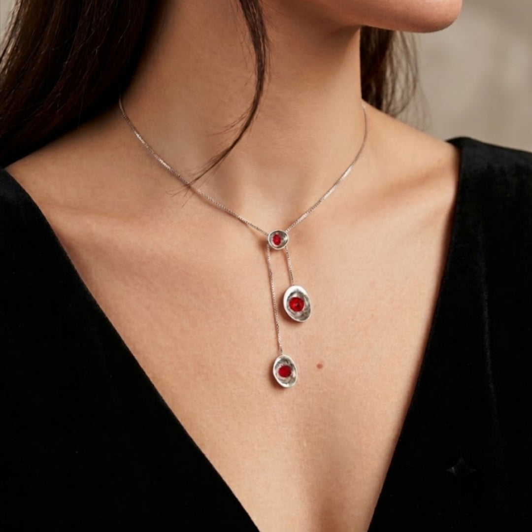 A silver lariat-style necklace with three dangling oval pendants, each featuring a textured silver border and a central red circular detail, shown worn on a model's neck on the left and hanging against a light stone background on the right. Made in Palestine. Brand